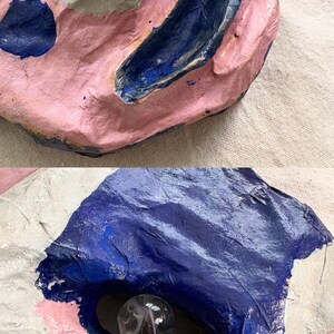 May include: Abstract sculpture with a textured surface. The artwork features pink, white, and blue paint. A clear glass marble is set into a dark blue recess. The piece has a handmade, organic appearance.