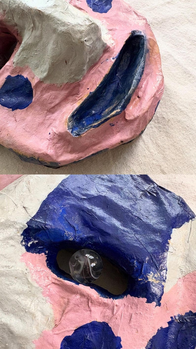 May include: Abstract sculpture with a textured surface. The artwork features pink, white, and blue paint. A clear glass marble is set into a dark blue recess. The piece has a handmade, organic appearance.