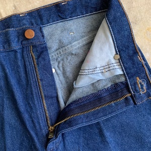 Puede incluir: Un par de pantalones vaqueros azules de mezclilla con cierre de cremallera y bot&oacute;n. La cremallera est&aacute; parcialmente abierta, revelando el interior de la cintura.