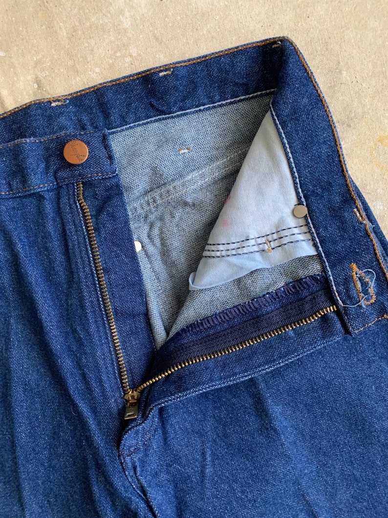 Puede incluir: Un par de pantalones vaqueros azules de mezclilla con cierre de cremallera y bot&oacute;n. La cremallera est&aacute; parcialmente abierta, revelando el interior de la cintura.