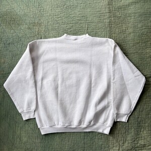 May include: A white crew neck sweatshirt with long sleeves. The sweatshirt is made of a soft material and is displayed on a green quilted surface. The garment has a ribbed hem and cuffs.