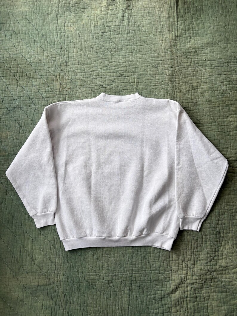 May include: A white crew neck sweatshirt with long sleeves. The sweatshirt is made of a soft material and is displayed on a green quilted surface. The garment has a ribbed hem and cuffs.