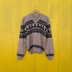 May include: A beige and brown knitted sweater with a V-neck and a geometric pattern of black llamas. The sweater is hanging on a hanger against a yellow wall.