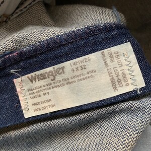 Puede incluir: Una etiqueta de cuidado blanca unida a un par de pantalones vaqueros de color azul oscuro. La etiqueta tiene el logotipo de Wrangler e instrucciones para lavar y cuidar los vaqueros. La etiqueta dice "Machine wash with like colors, only non-chlorine bleach when needed, tumble dry. Made in USA. 100% Cotton."
