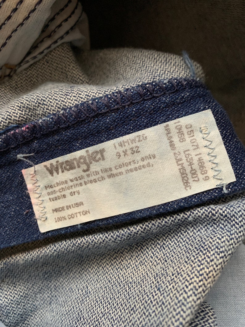 Puede incluir: Una etiqueta de cuidado blanca unida a un par de pantalones vaqueros de color azul oscuro. La etiqueta tiene el logotipo de Wrangler e instrucciones para lavar y cuidar los vaqueros. La etiqueta dice "Machine wash with like colors, only non-chlorine bleach when needed, tumble dry. Made in USA. 100% Cotton."