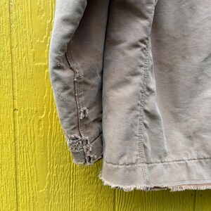 Peut inclure: Gros plan sur une manche de veste de travail beige us&eacute;e. Le tissu montre des signes d'usure, avec des bords effiloch&eacute;s et des zones ab&icirc;m&eacute;es. La veste est pos&eacute;e sur un fond en bois jaune vif.