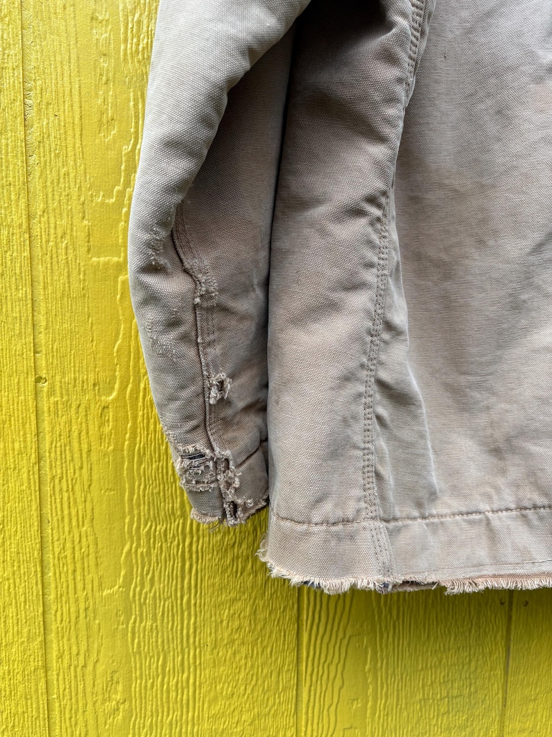 Peut inclure: Gros plan sur une manche de veste de travail beige us&eacute;e. Le tissu montre des signes d'usure, avec des bords effiloch&eacute;s et des zones ab&icirc;m&eacute;es. La veste est pos&eacute;e sur un fond en bois jaune vif.