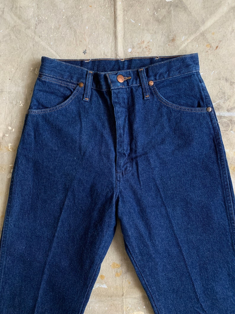 Puede incluir: Un par de pantalones vaqueros de mezclilla azul oscuro con un bot&oacute;n y remaches de cobre. Los pantalones tienen cintura alta y pierna recta.
