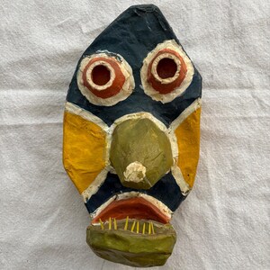 May include: A handmade, colourful mask with a stylised face. The mask features a dark blue background with yellow and white accents. The eyes are red with white outlines, and the nose is green. The mouth is red with yellow teeth.