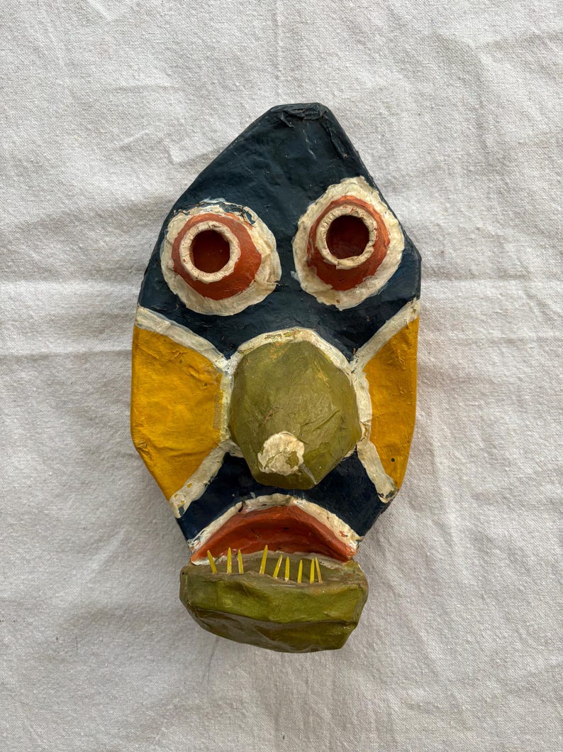 May include: A handmade, colourful mask with a stylised face. The mask features a dark blue background with yellow and white accents. The eyes are red with white outlines, and the nose is green. The mouth is red with yellow teeth.