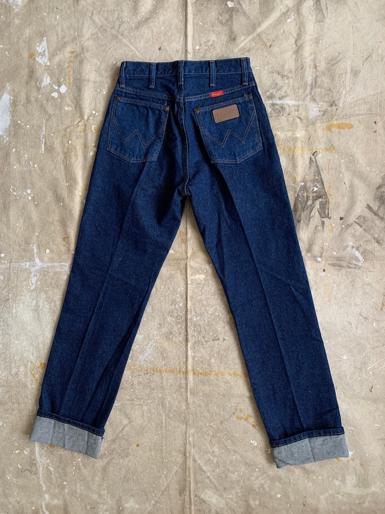 Puede incluir: Un par de pantalones vaqueros de mezclilla azul oscuro con un parche de cuero marr&oacute;n en el bolsillo trasero que dice "Wrangler". Los pantalones tienen un dobladillo con vuelta.