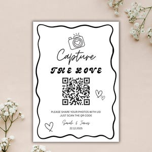May include: A white wedding sign with black text and a decorative border. The sign reads "Capture the Love" and includes a QR code, hearts, and the names Sarah & James with the date 22.12.2025. A camera icon is at the top.