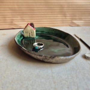 Könnte beinhalten: Handgefertigte Keramik-Ringschale mit dunkelgrünem Innenraum und einem kleinen Keramikhaus. Ein Silberring mit einem Türkisstein liegt in der Schale. Die Schale steht auf einer hellen Oberfläche.