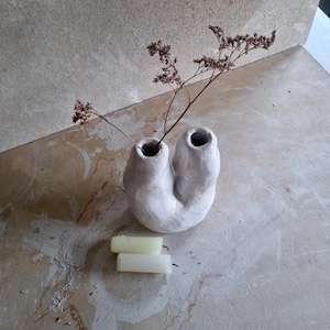 Könnte beinhalten: Eine weiße Keramikvase mit zwei Öffnungen, die getrocknete braune Stiele enthält. Zwei weiße Kerzen sind unter der Vase platziert. Die Vase hat eine einzigartige, gebogene Form. Der Hintergrund ist eine neutrale, strukturierte Oberfläche.