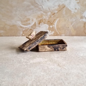 Könnte beinhalten: Eine kleine, rechteckige Keramikdose mit Deckel. Die Dose ist mit einer braunen Glasur verziert und hat eine kleine Vogelfigur auf dem Deckel. Der Vogel ist mit rosa und weißen Akzenten bemalt.