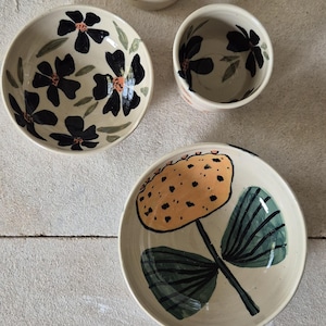 May include: A set of four ceramic vessels including two bowls and two cups. The pieces feature cream-colored surfaces with hand-painted designs of black flowers, green leaves, and a large yellow dotted flower with dark green foliage.