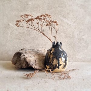 Könnte beinhalten: Eine kleine, keramikvase in Form eines Granatapfels. Die Vase ist in Schwarz und Weiß mit einem Marmoreffekt bemalt. Getrocknete Blumen befinden sich in der Vase.