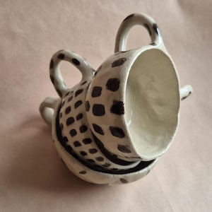 Könnte beinhalten: Ein Stapel handgefertigter Keramik-Tassen und Untertassen. Die Tassen und Untertassen sind cremefarben mit einem Muster aus dunkelbraunen Punkten. Jede Tasse hat einen gebogenen Henkel. Das Set steht auf einer hellbraunen Oberfläche.