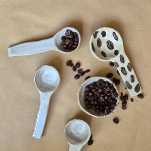 Könnte beinhalten: Handgefertigte Keramik-Löffel und eine kleine Schale, alle gefüllt mit dunklen Kaffeebohnen. Ein Löffel hat ein gepunktetes Design. Die Artikel sind auf einer braunen Oberfläche angeordnet und erzeugen eine rustikale Ästhetik.