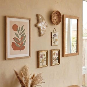 Könnte beinhalten: Eine Wanddekoration mit gerahmter Kunst, einem Spiegel und dekorativen Objekten. Die Kunst umfasst botanische Drucke und ein abstraktes Design. Ein Holztisch beherbergt eine Vase mit getrockneten Pflanzen und ein Buch. Die Farbpalette ist neutral mit warmen Tönen.