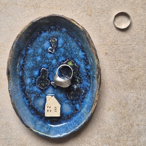 Könnte beinhalten: Eine blaue und braune Keramikschale mit strukturierter Oberfläche, die einen Silberring und eine kleine weiße Hausfigur enthält. Ein zweiter Silberring liegt auf der Oberfläche neben der Schale.