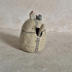 Könnte beinhalten: Ein gesprenkeltes beiges Keramikgefäß mit Deckel, verziert mit einem kleinen skulptierten Haus auf der Oberseite und einem handgemalten schwarzen Leitermotiv, das vom Deckel bis zum Boden reicht. Ein passender Keramiklöffel steckt im Gefäß.