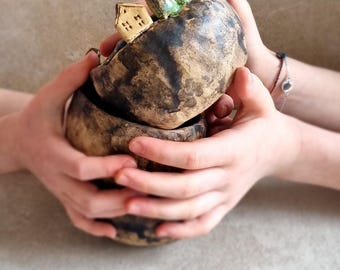 May include: A handmade ceramic bowl with a rustic, textured finish. The bowl contains a small, detailed house figurine and decorative elements. The bowl is held by two hands against a neutral background.