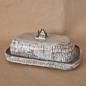 May include: A ceramic butter dish with a lid, featuring a textured, off-white and brown design. The lid is topped with a small, decorative house-shaped handle. The dish is rectangular with a matching base.