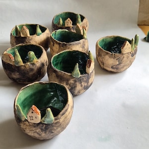 Op de afbeelding: Een verzameling van zes kleine, ronde keramische kommen met een getextureerde, beige buitenkant en een glanzende, diepgroene binnenkant. Elke kom is voorzien van miniatuur handgevormde kleihuisjes en groene bomen aan de rand.