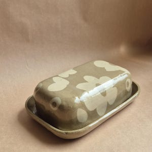 Könnte beinhalten: Ein Keramik-Butterdose mit Deckel, mit einer beigefarbenen Grundfarbe und einem Muster aus weißen Blumenformen. Die Dose ist rechteckig mit abgerundeten Kanten und steht auf einem passenden Tablett.