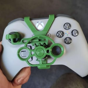 Op de afbeelding: Een groene 3D-geprinte bevestiging voor een Xbox-controller. De bevestiging heeft een ronde grip met een tandwielontwerp en een duimsteun. De bevestiging is bevestigd aan de rechterkant van de controller, in de buurt van de ABXY-knoppen.