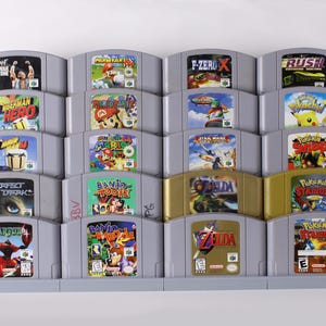 May include: A collection of Nintendo 64 video game cartridges, featuring titles like "Mario Kart 64", "The Legend of Zelda", and "Pokémon Stadium". The cartridges are arranged in a gray display rack, showcasing the colorful game box art.