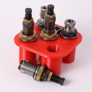 May include: A red plastic holder with several metal and brass components. The components have a variety of shapes and sizes, with threaded sections and circular bases. One component rests outside the holder.