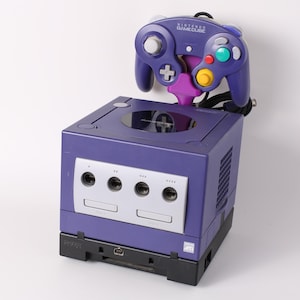 May include: A purple Nintendo GameCube console with a matching controller. The console has a silver faceplate with four ports and a disc tray. The controller is connected and features various colored buttons.