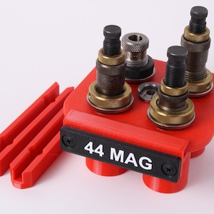 May include: A red 3D-printed reloading tool holder with multiple metal components. The holder has a black label that reads "44 MAG". Several metal pieces are mounted on a red base, and there are also red, parallel, grooved pieces.