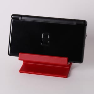 May include: A black handheld gaming console is displayed on a bright red stand. The console has a glossy finish and a simple design. The stand is a solid, rectangular shape, providing a stable base for the device. The background is plain white.