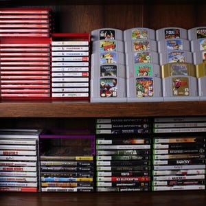 May include: A wooden shelf filled with video game cartridges and cases. The top shelf displays stacks of red game cases, gray Nintendo 64 cartridges, and a yellow and white Pokeball. The lower shelves hold various game cases.