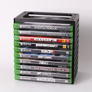 May include: A black plastic game organizer holding multiple Xbox One video game cases. The cases have green spines and various game titles visible, including "Shadow of Mordor" and "Red Dead Redemption II".