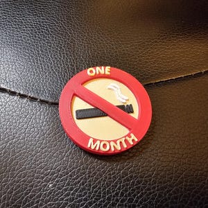 May include: A red, white, and black circular emblem with the words "ONE MONTH" and a no-smoking symbol. The emblem is on a textured black surface.