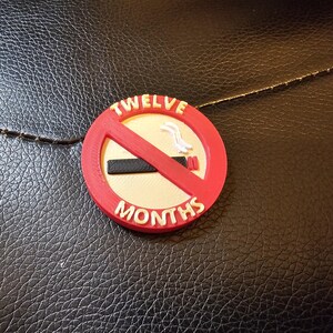 May include: A round, red and white "No Smoking" emblem with the words "TWELVE MONTHS" in white. The emblem features a red slash through a black cigarette. The background is a textured black surface.