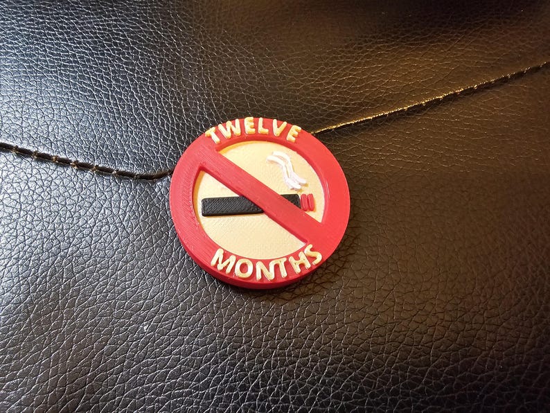 May include: A round, red and white "No Smoking" emblem with the words "TWELVE MONTHS" in white. The emblem features a red slash through a black cigarette. The background is a textured black surface.