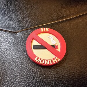 May include: A round, red and white "No Smoking" sign with the text "SIX MONTHS". The sign has a red slash through a cigarette. The sign is on a textured black surface.