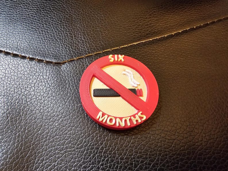 May include: A round, red and white "No Smoking" sign with the text "SIX MONTHS". The sign has a red slash through a cigarette. The sign is on a textured black surface.