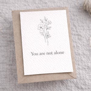 May include: A white card with a floral illustration and the text "You are not alone" is placed on a brown envelope. The card and envelope are on a textured, light gray surface. A ball of twine is in the background.