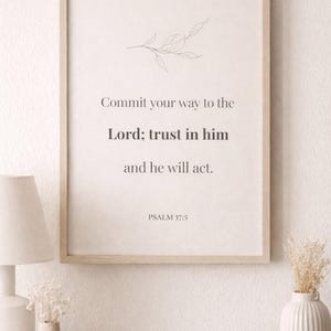 May include: A framed print with the text "Commit your way to the Lord; trust in him and he will act. Psalm 37:5." The print has a light wood frame and a minimalist botanical illustration. The print is displayed on a white wall.