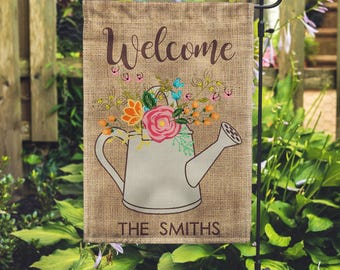 May include: A garden flag with a burlap texture features a watering can design filled with colorful flowers. The word "Welcome" is written at the top in a script font, and "THE SMITHS" is printed at the bottom.
