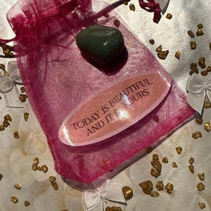 May include: A small, sheer magenta gift bag with a drawstring closure. Inside, a smooth, green stone and a pink oval with the text "TODAY IS BEAUTIFUL AND IT OURS". The background has gold flecks and white bows.