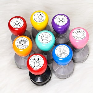May include: A collection of colorful, round stamps with gray bases. Each stamp has a different design and message, such as "You are a star!" and "Thumbs up for effort!" The stamps are in various colors, including red, yellow, and blue.