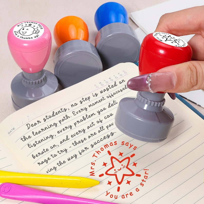 May include: A collection of colorful teacher stamps with gray bases. One stamp is being used to stamp a notebook. The stamps have different colored tops: pink, orange, blue, and red. The stamp says "Mrs. Thomas says You are a star!"