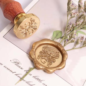 May include: A gold-coloured wax seal stamp with a wooden handle and a floral design with the letter "G". A matching wax seal is also present. The stamp and seal are on a white surface with text.